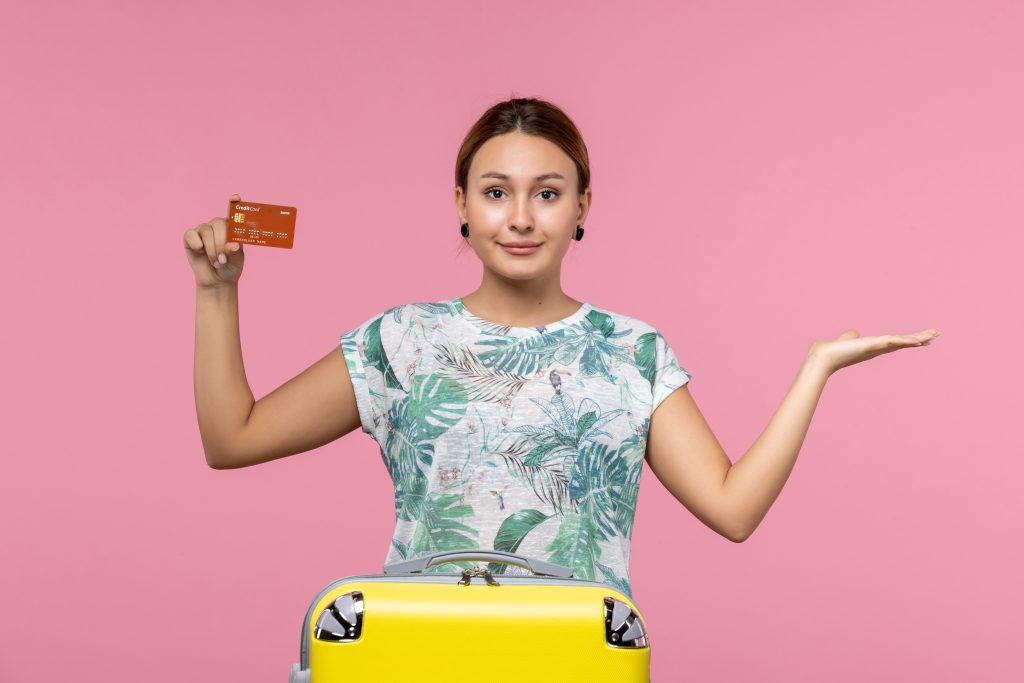 A woman holding up a credit card, standing near a suitcase.